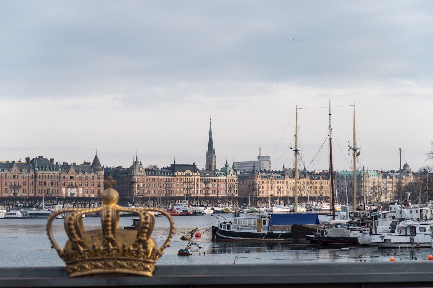 Im Vordergrund eine Krone, im Hintergrund hanseatische große Häuser, dazwischen Segelboote auf dem Wasser